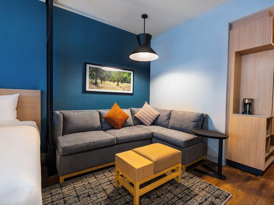 A modern room with a grey sofa, a wooden side table, a small wooden bench, a framed picture on the wall, and a wardrobe.