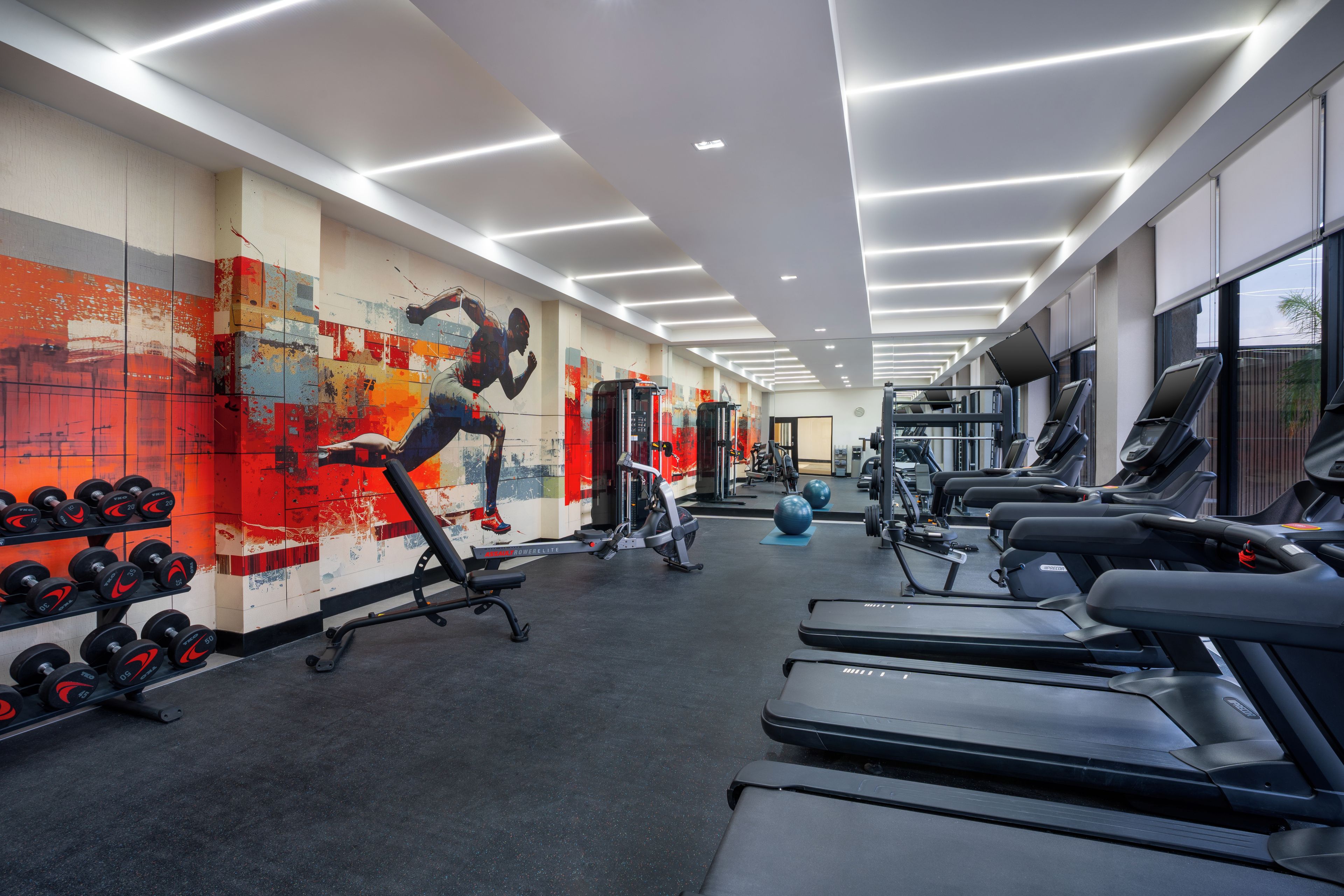 Un gimnasio con cintas de correr, máquinas de pesas y una pared con un mural colorido.