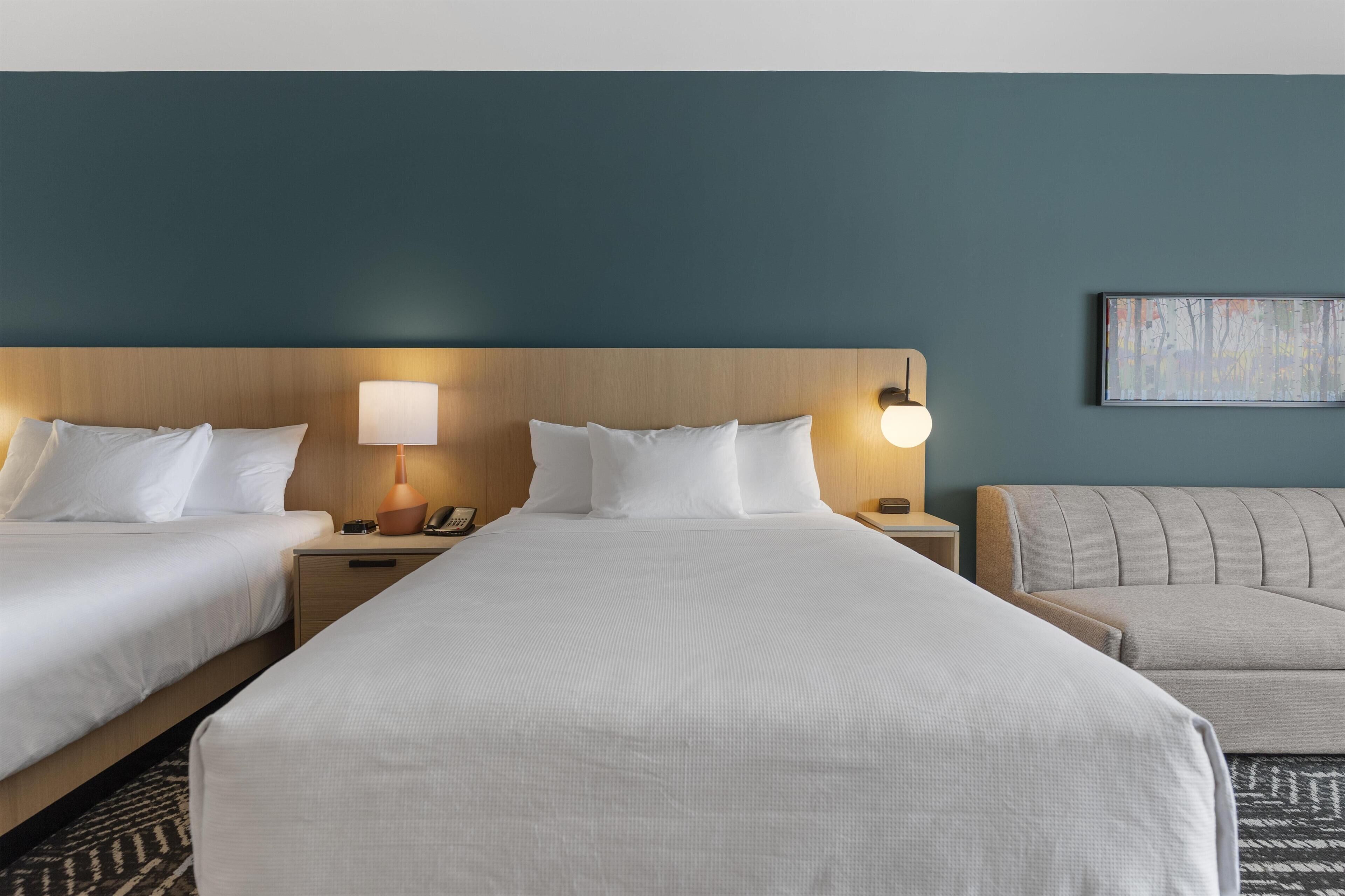 Une chambre d’hôtel avec deux lits, un canapé, une table de chevet avec une lampe, un téléphone et un tableau au mur.