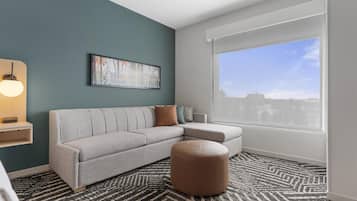 A modern living room with a grey sectional sofa, a brown ottoman, a large window with blinds, a framed abstract painting, and a pendant light.