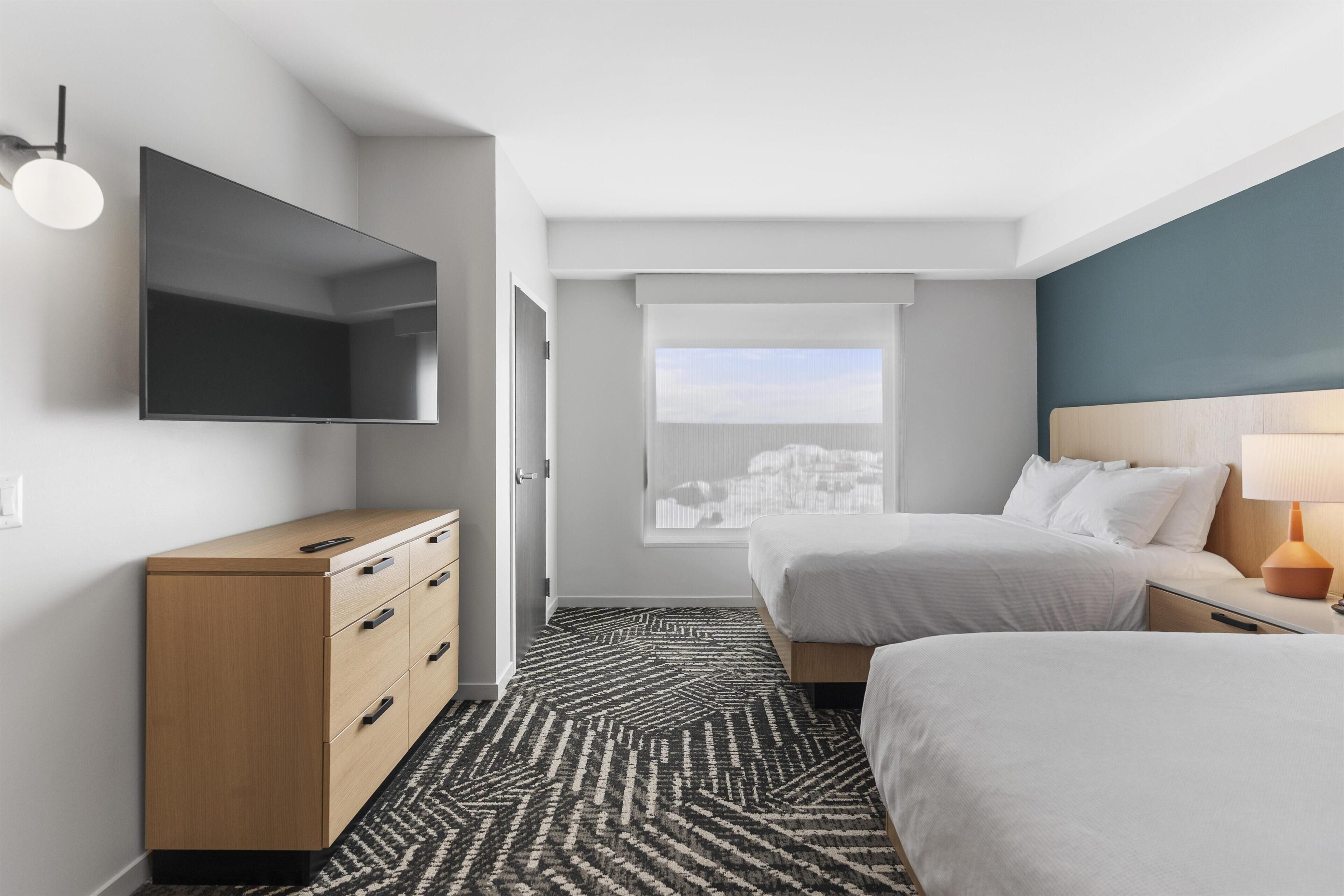 Habitación de hotel con cama, mesita de noche, lámpara, televisor, armario y ventana con vistas.