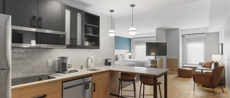 A modern kitchen with a dishwasher, a coffee machine, and a microwave. There is a dining area with a table and chairs, and a living room with a TV and seating.