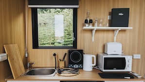 Een compact keukenhoekje met een spoelbak, magnetron en koffiezetapparaat, een raam met uitzicht op groen, en een plank met keukengerei.