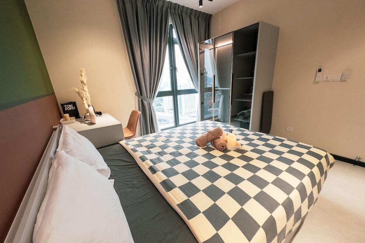 A modern hotel room with a checkered bedspread, a large window with curtains, a wardrobe, and a small table with a vase and a sign.