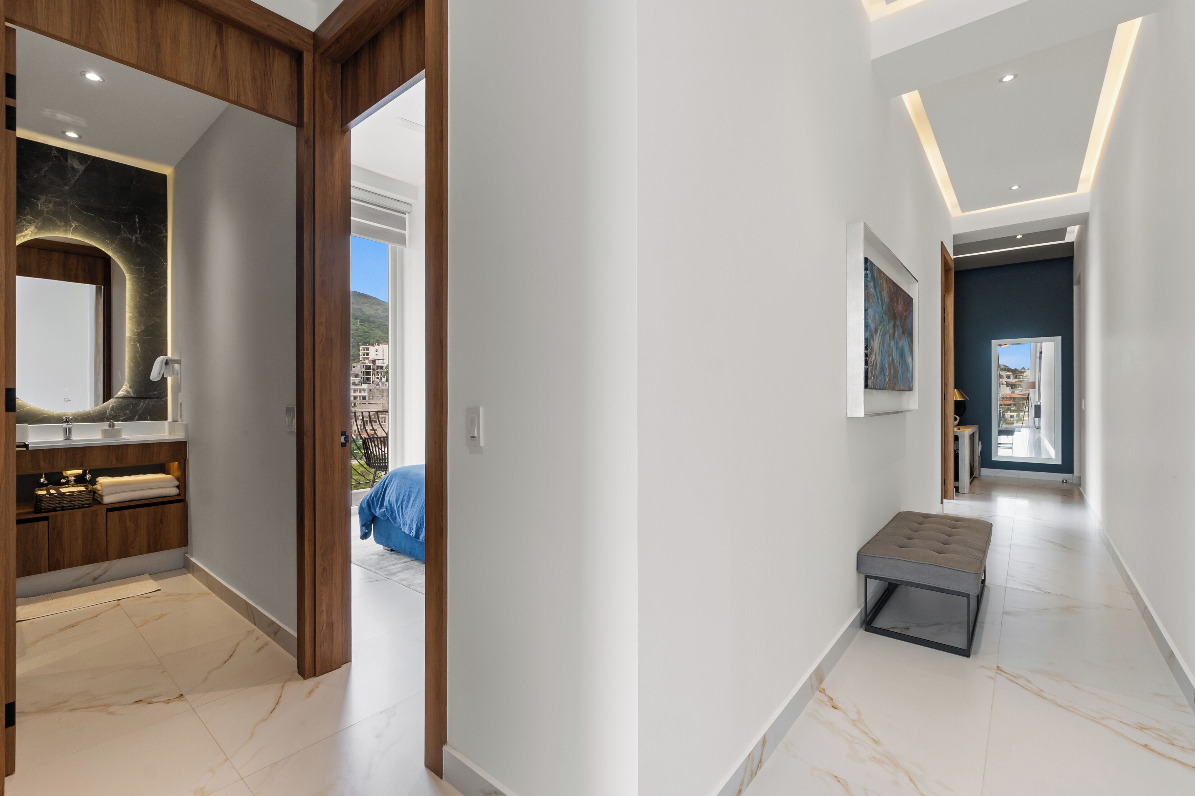 A modern hallway with a wooden door leading to a bathroom, a bench, and a view of the outside.