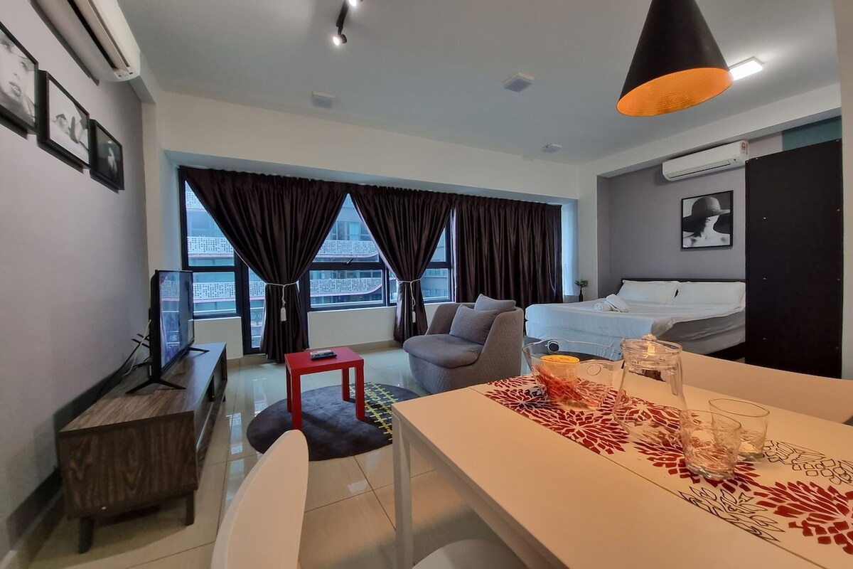A modern hotel room with a large window, a bed, a desk with a computer, a red chair, and a dining table with glasses.