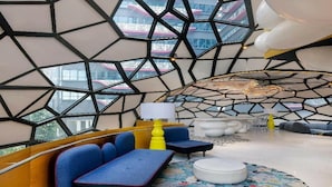 Un interior modern amb patrons geomètrics a la paret, un sofà blau, un otomà rodó i una làmpada groga.