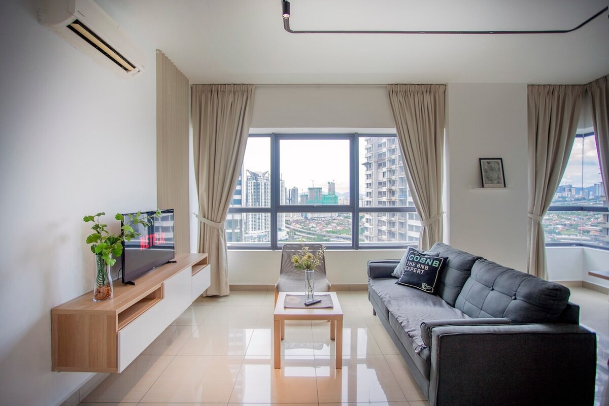 A modern living room with a flat-screen TV, a grey sofa, a coffee table, and a view of a cityscape through large windows.
