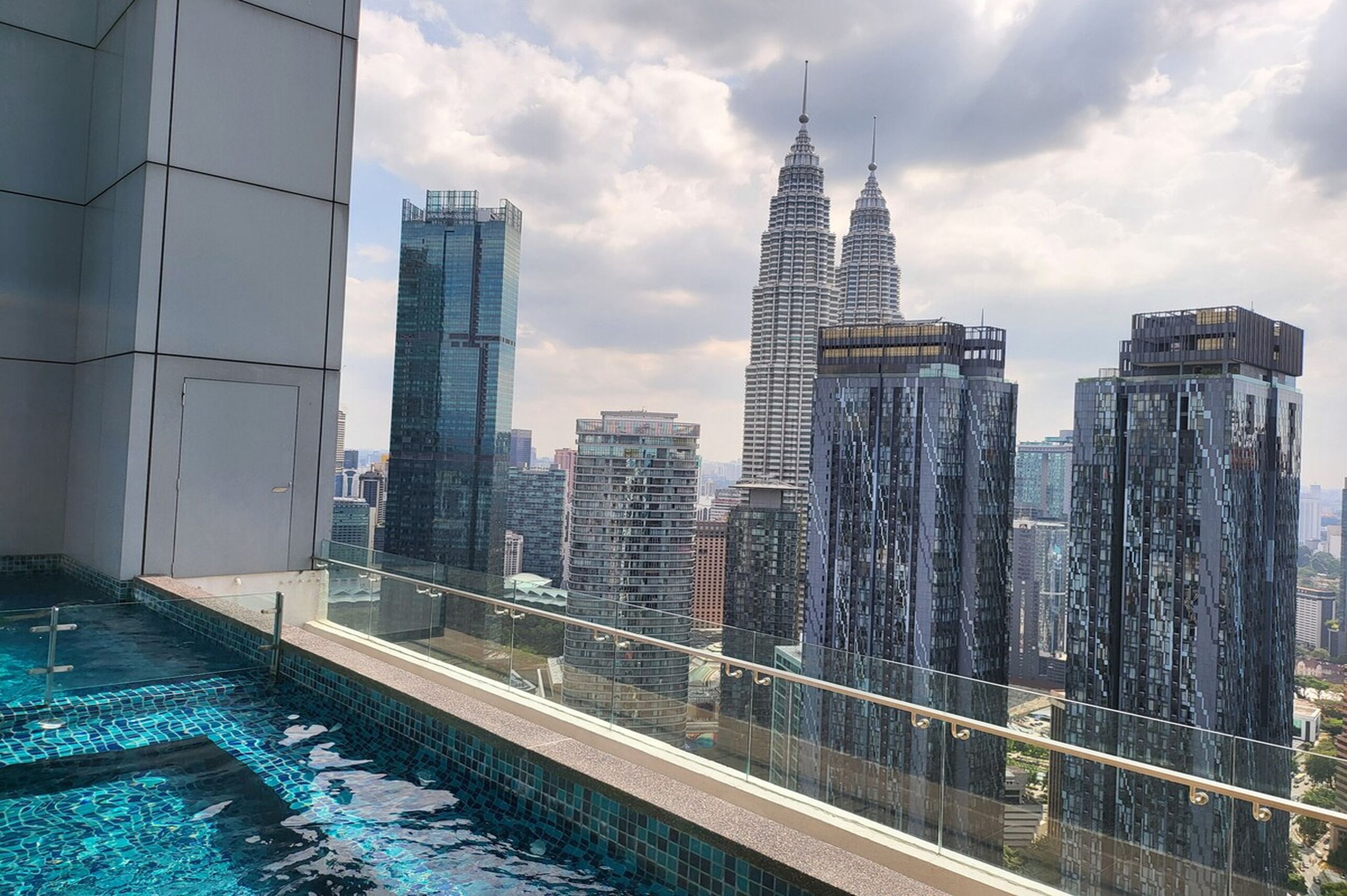 A high-rise building with a glass balcony overlooking a cityscape, including the Petronas Towers.