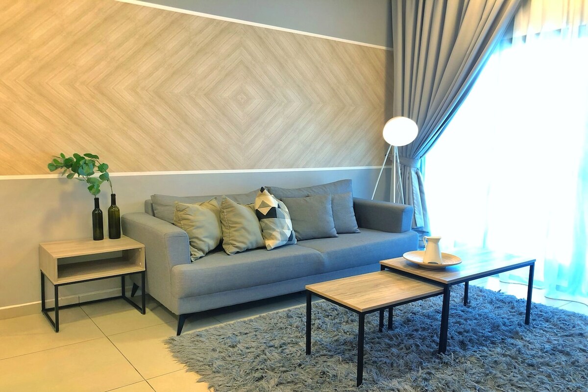 A modern living room with a grey sofa, a small wooden coffee table, a side table with two bottles, a plant, and a floor lamp.