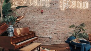A cozy room with a grand piano, a neon sign reading "Star Piano", and a brick wall. - Starr Piano: Roxie Suite –  Walk to Broadway! (Nashville)