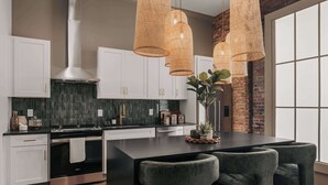 A modern kitchen with white cabinets, a black countertop, and a large island with chairs. There are wicker pendant lights and a brick wall in the background. - The Carraway Suite • Luxury • Walk to Broadway! (Nashville)