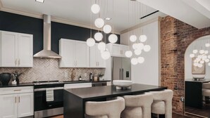 Una cocina moderna con armarios blancos, una isla central con taburetes de bar, una campana extractora de acero inoxidable y una lámpara colgante.