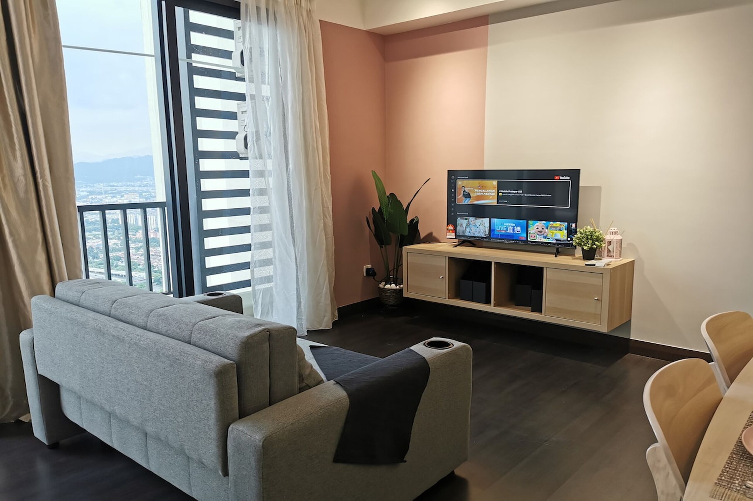 Una sala de estar moderna con un sofá gris, un mueble de televisión de madera y vistas a la ciudad a través de ventanas amplias.