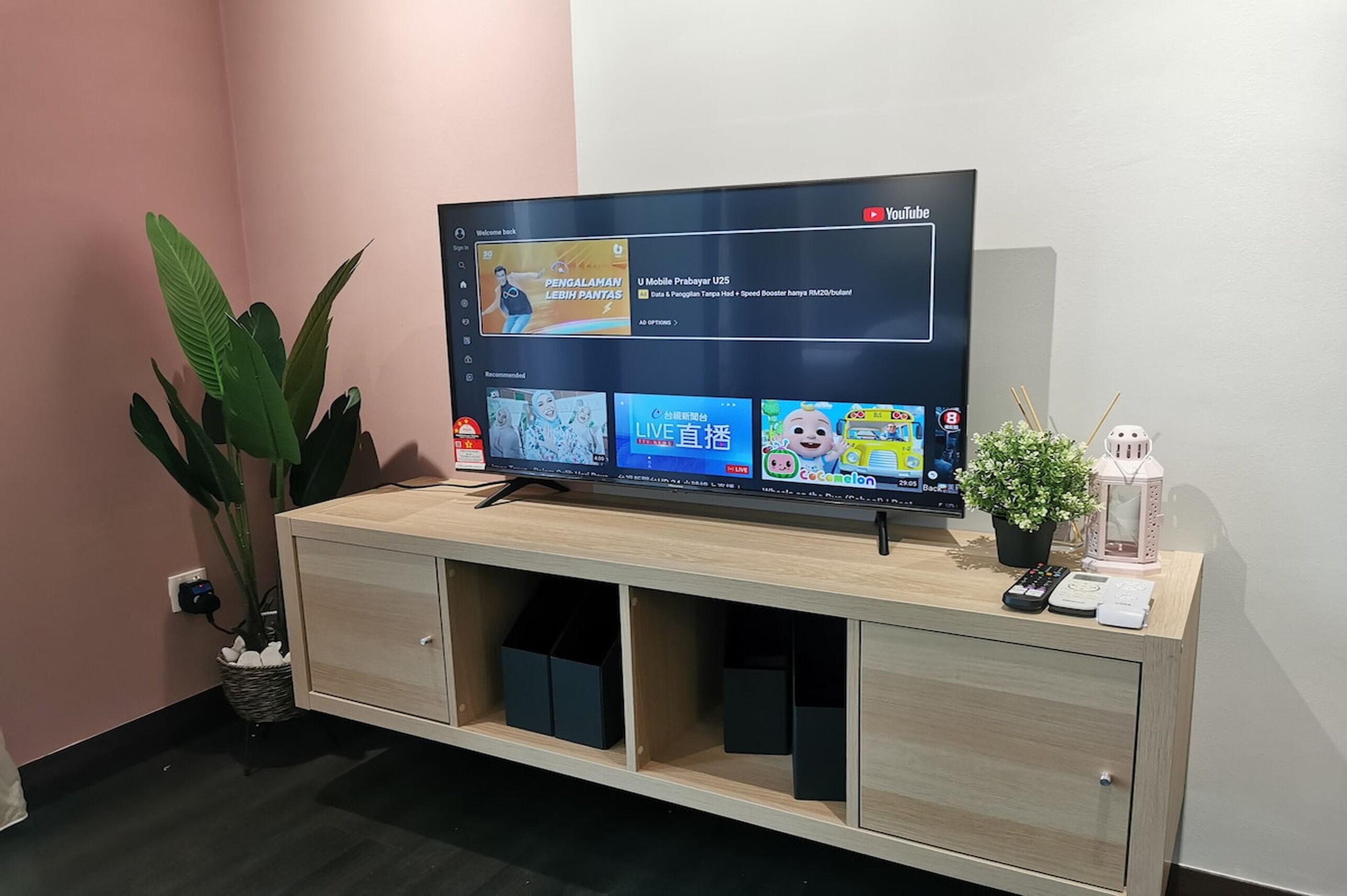 Una TV a schermo piatto su un mobiletto in legno con interfaccia YouTube, una pianta in vaso e un tavolino con un telecomando e un telefono.
