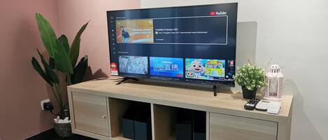 Una TV a schermo piatto su un mobiletto in legno con interfaccia YouTube, una pianta in vaso e un tavolino con un telecomando e un telefono.