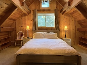 Un dormitorio de cabaña de madera con una cama, dos mesitas de noche, una silla y una ventana.
