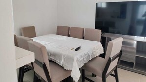 A dining area with a table covered by a white tablecloth, surrounded by beige chairs. A flat-screen TV is mounted on the wall to the right.