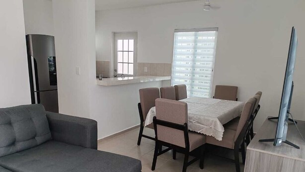 A modern living room with a grey sofa, a dining table with chairs, a TV on a stand, and a kitchen area in the background.
