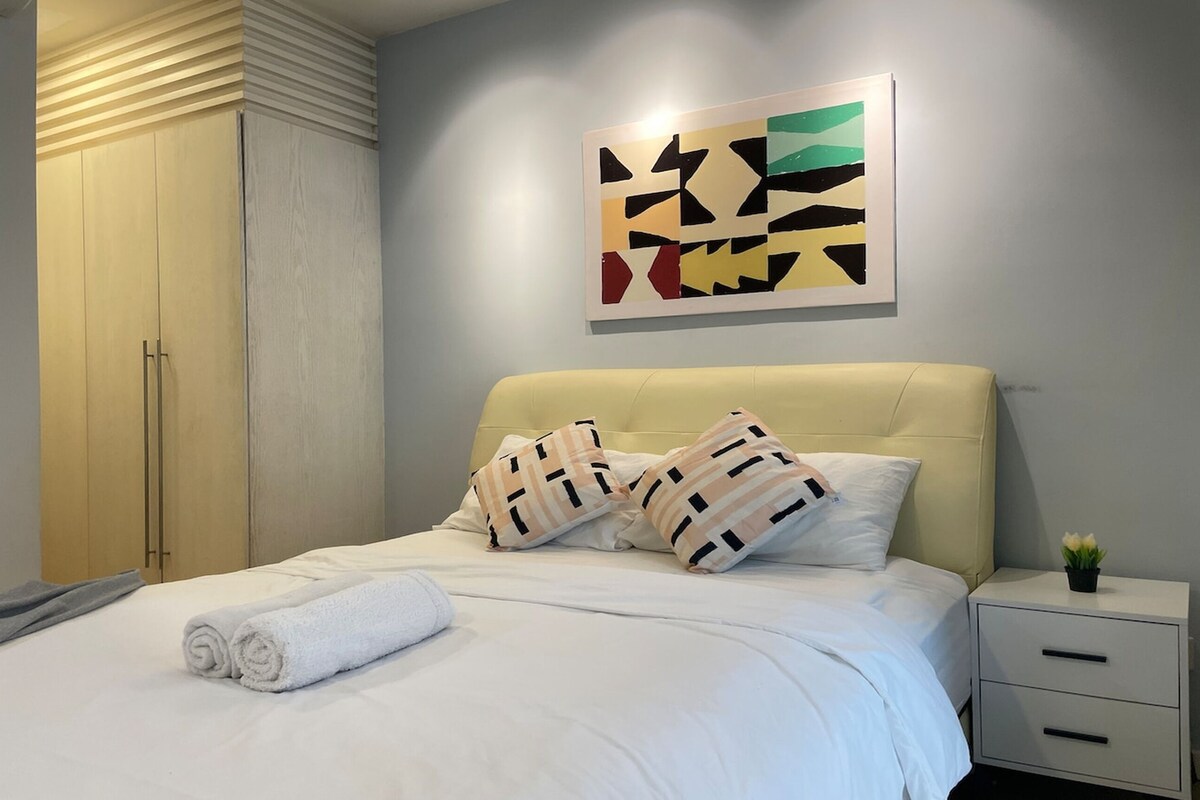 A neatly made bed with white linens and two decorative pillows, a bedside table with a small plant, and a wardrobe with a light wood finish.