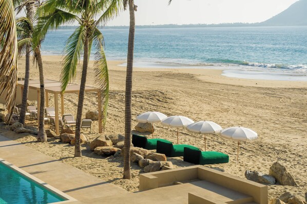 Un espace piscine situé au bord de la plage, avec des chaises longues, des parasols et des palmiers.