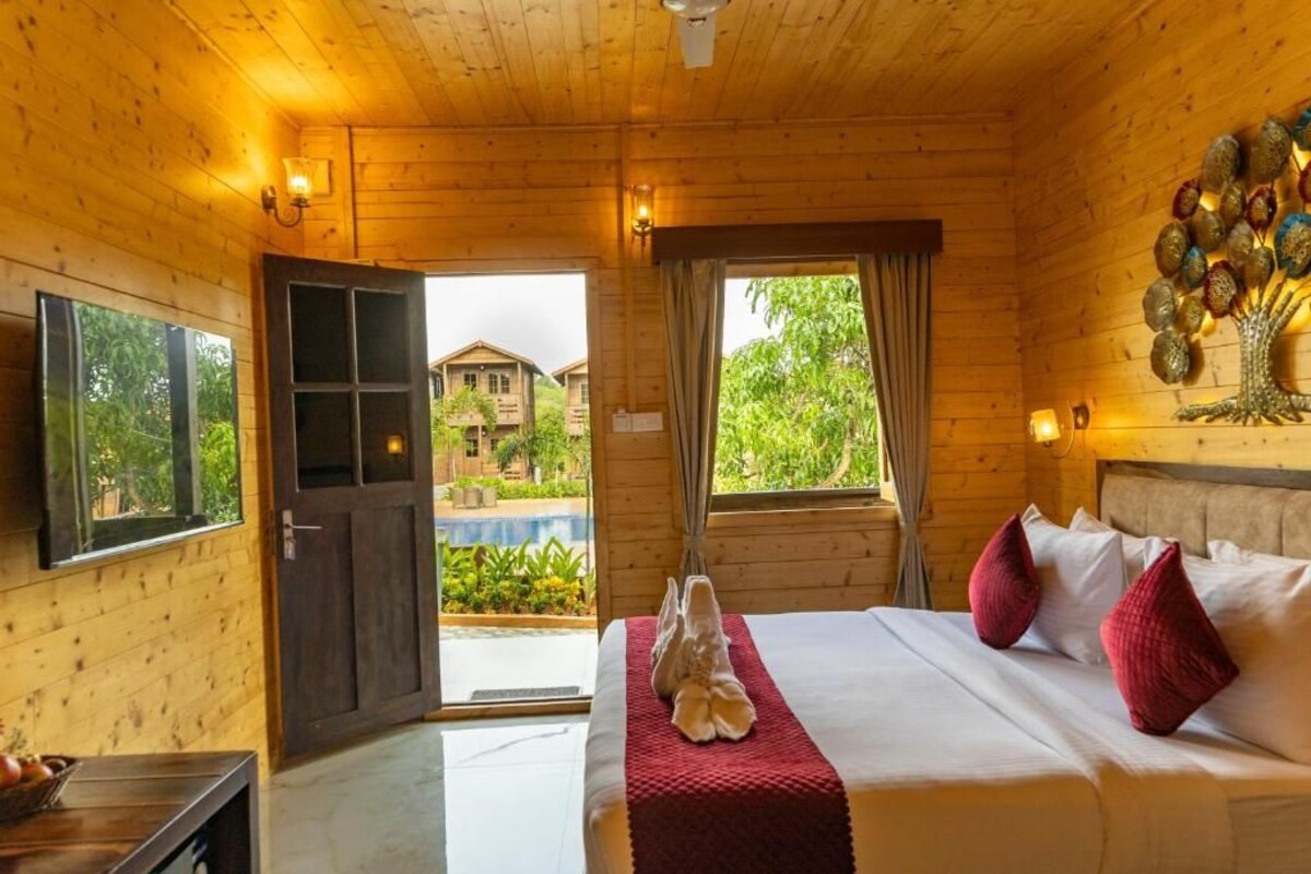 A wooden cabin room with a bed, a window, a door, and a view of a house and greenery outside.