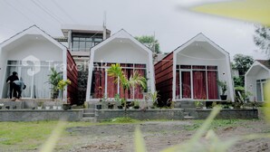 Une rangée de maisons blanches, de plain-pied, avec des rideaux rouges, entourées de verdure et bordées d’un sol en terre.