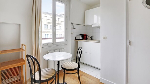 A small, bright kitchen with white cabinets, a round table, two chairs, and a window with curtains. - A Canal St Martin Lovely Room (Paris)