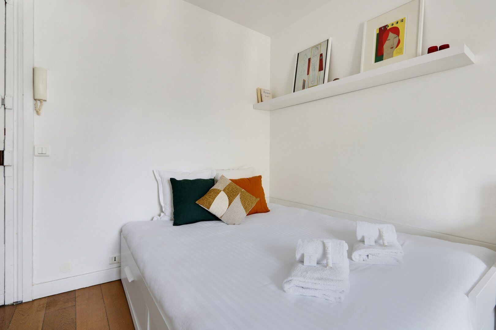 Un lit bien fait, avec du linge de lit blanc et deux serviettes, un oreiller vert et beige, et une petite étagère avec des photos encadrées.
