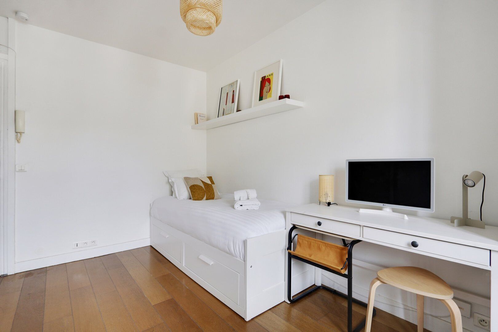 Et minimalistisk soveværelse med seng, skrivebord, stol, fjernsyn og reol med bøger og billeder.