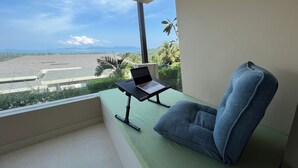 Un balcon avec un ordinateur portable posé sur un support, une chaise bleue et une vue sur une plage et des montagnes.