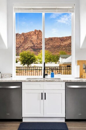 Una cocina con armarios blancos, un lavavajillas de acero inoxidable y un ventanal con vistas a un paisaje rocoso.