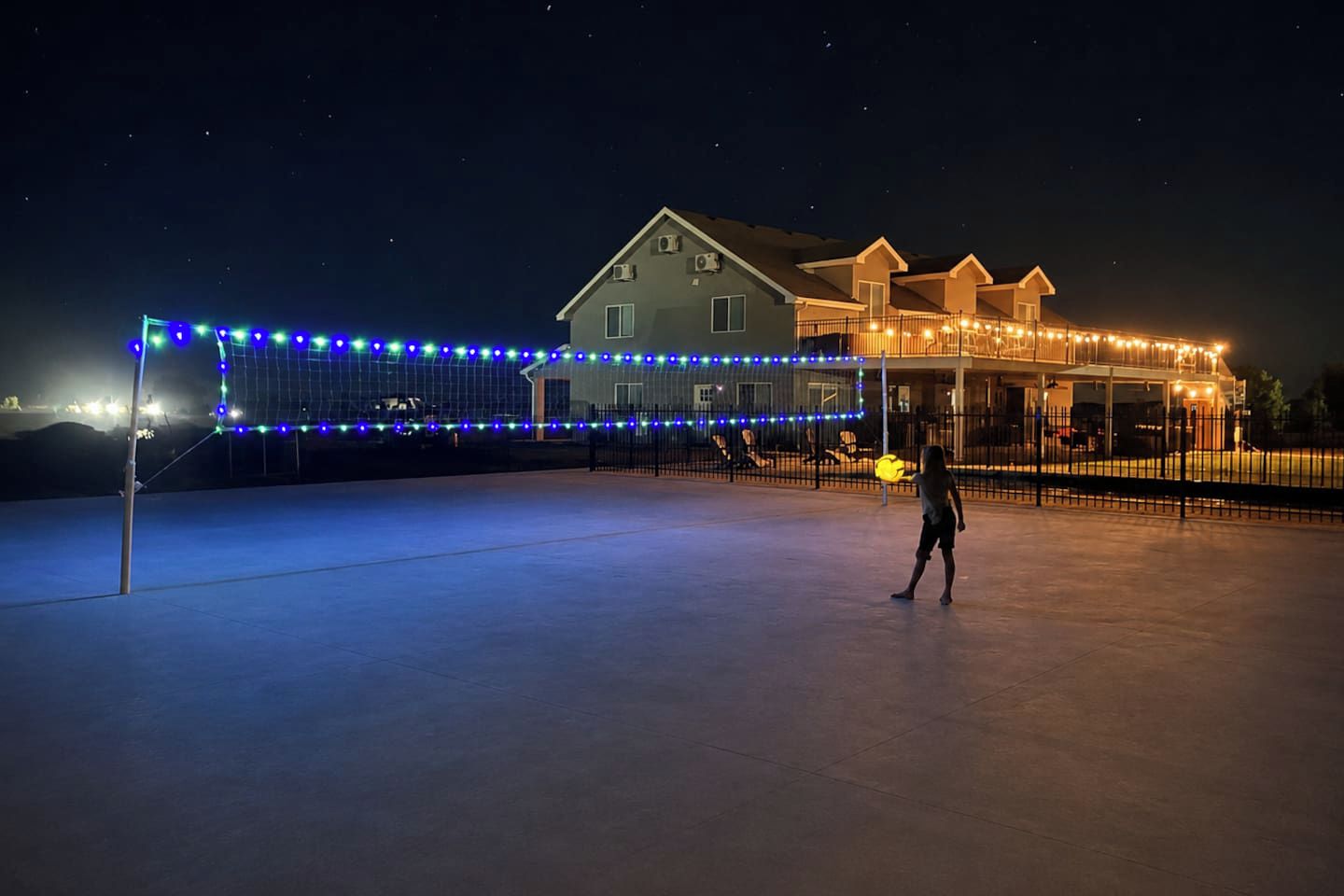 An outdoor tennis court with a net, surrounded by a fence and illuminated by string lights.