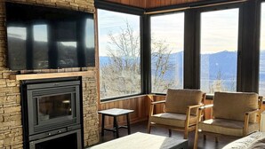 Ein Zimmer mit Kamin aus Stein, Holzmöbeln und großen Fenstern mit Blick auf die Berge.