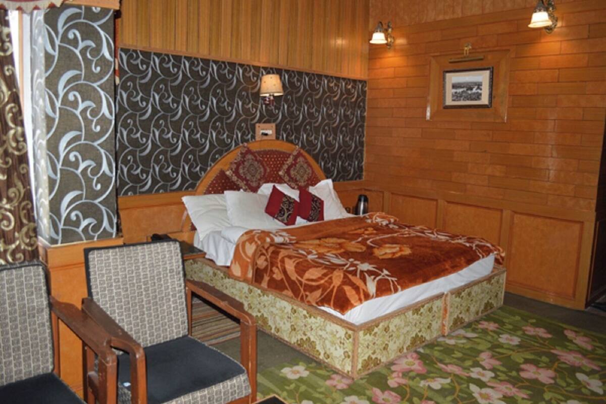 A bedroom with a wooden headboard, a floral bedspread, and a patterned wall. There is a chair with a cushion and a lamp on the wall.