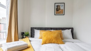 A neatly made bed with white linens and a mustard-colored throw blanket, a framed artwork on the wall, and a window with curtains. - Cozy 4 bedroom in Aintree -Liverpool Free Parking, Super fast Wifi Internet. (Bootle)