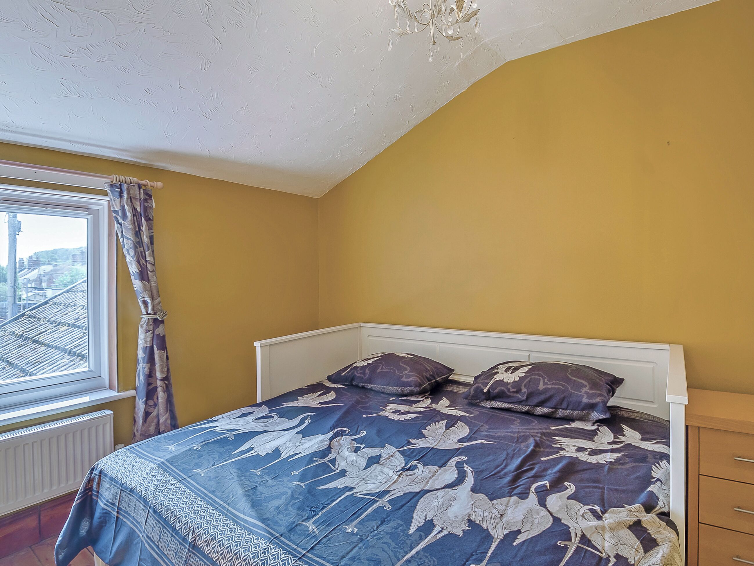 A bedroom with a bed, a window with curtains, a radiator, and a wooden cabinet.