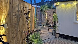 A courtyard with a wooden fence, a brick wall, and a white building. There are potted plants and a metal chair. - 3 bedroom accommodation in Beccles (Beccles)