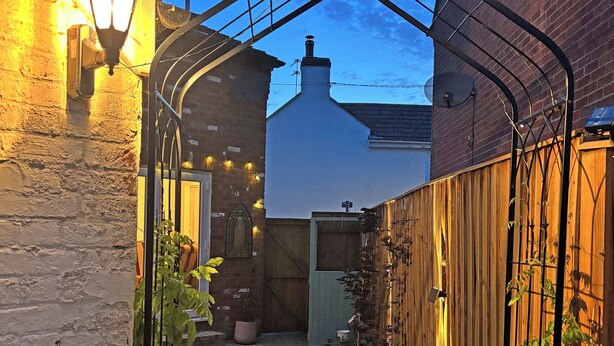 A narrow alleyway with a brick building on the left and a wooden fence on the right. There are hanging lights and a small garden area with plants. - 3 bedroom accommodation in Beccles (Beccles)