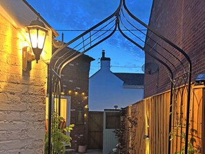 Une ruelle étroite, avec un bâtiment en briques sur la gauche et une clôture en bois sur la droite. Des lampes sont suspendues et il y a un petit espace jardin avec des plantes.
