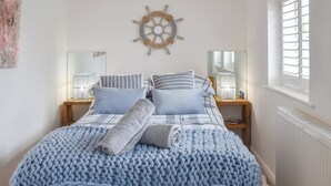 Una cama bien hecha con una manta y almohadas de punto azul, cabecera de madera, decoración con timón de barco y una ventana con persianas.