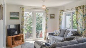 A living room with a television on a wooden stand, a grey sofa, and a window with curtains. - A spacious holiday retreat with private, enclosed garden, nestled within St. (Padstow)