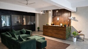 A modern reception area with a wooden desk, a green sofa, and a ceiling fan. - Hotel Stayzzy Bloom (Noida)