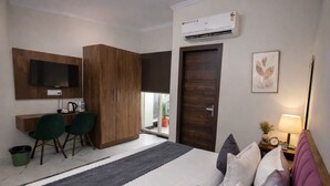 A bedroom with a bed, a desk, two chairs, a wardrobe, an air conditioner, and a window with a view. - Hotel Stayzzy Bloom (Noida)