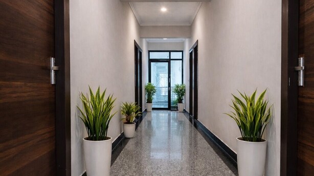 A hallway with dark wooden doors, white walls, and a polished granite floor. There are potted plants on either side of the hallway. - Hotel Stayzzy Bloom (Noida)