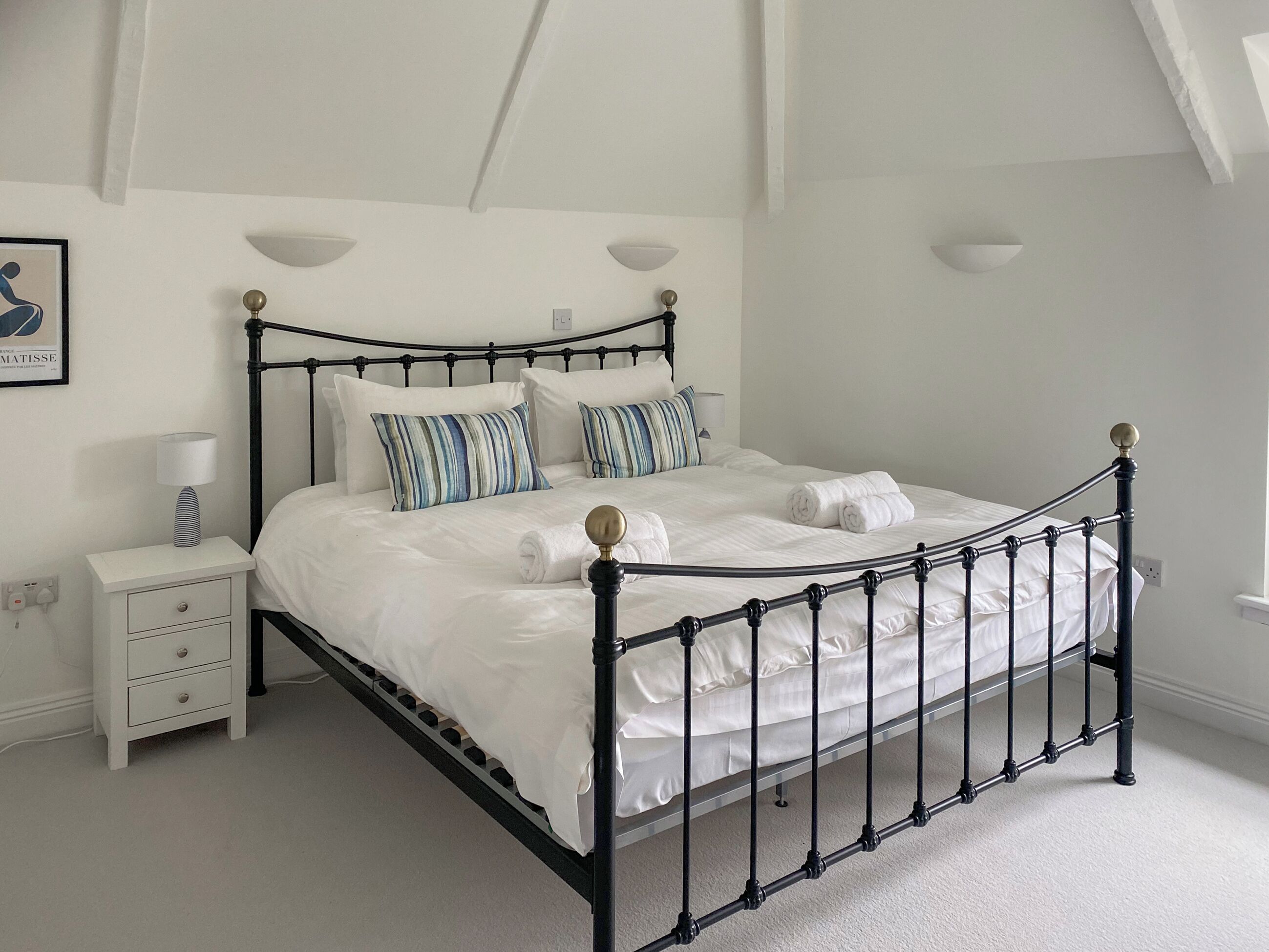 A neatly made bed with a black metal frame, white bedding, and striped pillows. There is a bedside table with a lamp and a drawer. The room has white walls and a sloped ceiling.