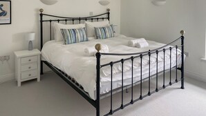 A neatly made bed with a black metal frame, white bedding, and striped pillows. There is a bedside table with a lamp and a drawer. The room has white walls and a sloped ceiling. - 3 bedroom accommodation in St Mawes (St Mawes)