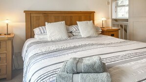 A neatly made bed with striped bedding and a folded towel, wooden headboard, bedside lamps, and a window with blinds. - 3 bedroom accommodation in Bude (Bude)