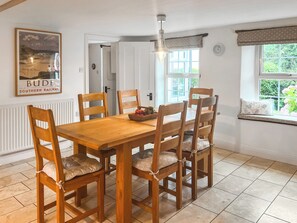A dining area with a wooden table and chairs, a fruit bowl, a framed picture on the wall, and a window with a view of greenery.