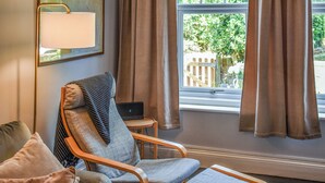 A room with a wooden armchair, a small table with a laptop, and a view of greenery outside.